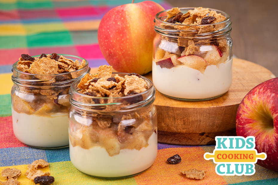 3 parfaits made with yogurt, cereal and apples in glass jars on a colorful table cloth- 2 apples are beside the glass jars.