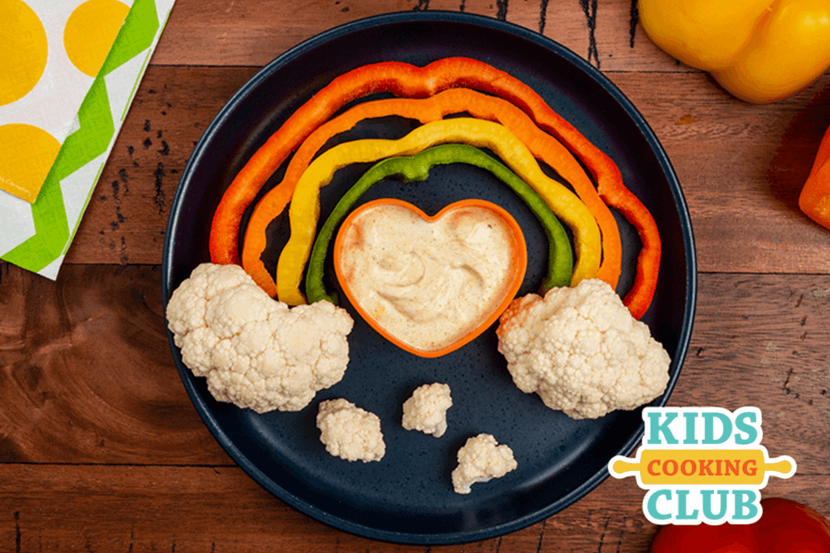rainbow made with colorful peppers and cauliflower as clouds with a heart-shaped bowl filled with dip
